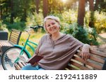Small photo of smiling modern old lady relaxing city park. Pensive senior gray haired woman casual sitting bench outdoors reading book. cycling forest park, bicycle, healthy active lifestyle after 50-60 years