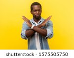 Never again. Portrait of absolutely convinced man in denim casual shirt standing crossing hands, showing x sign meaning stop, there is no way, finish. indoor studio shot isolated on yellow background