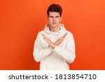 There is no way! Serious concentrated young man in stylish hoodie showing no gesture crossing arms, say no to bad habits, addiction. Indoor studio shot isolated on orange background