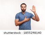 Small photo of I swear! Conscious honest man raising one hand and holding on chest another, swearing in honesty and devotion, patriotism. Indoor studio shot isolated on white background