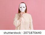 Portrait of unknown anonymous woman covering her face with white mask, hiding her real personality, anonymity, wearing white sweater. Indoor studio shot isolated on pink background.