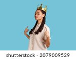 Small photo of Portrait of proud confident arrogant woman with black dreadlocks, standing in gold crown, pointing at herself with pride, wearing white shirt. Indoor studio shot isolated on blue background.