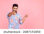 Small photo of Young adult serious angry man pointing at you with finger and presenting area with promotion, necessarily pay attention, has strict expression. Indoor studio shot isolated on pink background.