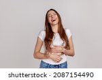 Small photo of Extremely happy woman holding her stomach and laughing out loud, chuckling giggling at amusing anecdote, sincere emotion, wearing white T-shirt. Indoor studio shot isolated on gray background.