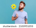 Small photo of Portrait of funny positive childish handsome bearded man wearing T-shirt playing blowing yellow windmill, having fun with paper toy. Indoor studio shot isolated on blue background.