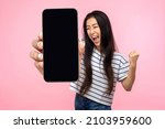 Small photo of Portrait of euphoric happy joyful girl with long hair screaming with happiness showing mobile advertisement mockup area and celebrating her victory. indoor studio shot isolated on pink background