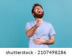 Hilarious laughter. Portrait of joyful happy bearded man laughing loudly and pointing to camera, mocking taunting you, holding belly. Indoor studio shot isolated on blue background.