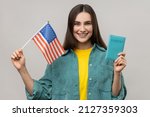 Young adult attractive woman holding united states flag and passport, being happy to move abroad to USA, wearing casual style jacket. Indoor studio shot isolated on gray background.