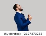 Side view of bearded man showing yes gesture and screaming celebrating his victory, success, dreams comes true, euphoria, wearing official style suit. Indoor studio shot isolated on gray background.
