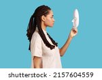 Side view of confident serious woman with dreadlocks holding and looking at white mask with attentive look, trying to understand hiding personality. Indoor studio shot isolated on blue background.