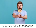 Portrait of excited amazed hungry handsome bearded man holding out sweet sugary confectionery, feels hungry, enjoying tasty lollipop. Indoor studio shot isolated on blue background.