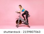 Small photo of Side view full length of beautiful sportswoman cycling a bike at home, cardio training, exercising legs, wearing sports tights and top. Indoor studio shot isolated on pink background.