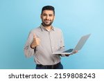 Small photo of Portrait of smiling positive businessman standing with portable computer in hand, looking at display, showing thumb up, wearing striped shirt. Indoor studio shot isolated on blue background.