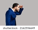 Side view of serious concentrated bearded man watching far away with hand above eyes, looking forward to future, wearing official style suit. Indoor studio shot isolated on gray background.