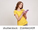 Portrait of glad curly haired teenage girl in yellow casual style T-shirt writing in notebook, filling diary or schedule, smiling sincerely. Indoor studio shot isolated on gray background.