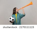 Side view of excited woman blowing in horn holding black and white soccer ball, celebrating victory of favourite football team, wearing casual jacket. Indoor studio shot isolated on gray background.