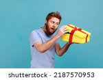 Portrait of cunning handsome bearded man unboxing yellow gift box, looking inside with curious facial expression, birthday present. Indoor studio shot isolated on blue background.