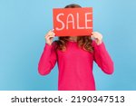 Portrait of unknown woman wearing pink pullover, covering face with Sale inscription, market discounts, black friday, low prices. Indoor her studio shot isolated on blue background.