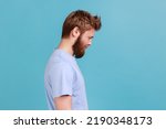 Side view of bearded upset young man standing and looking at camera with dissatisfied sadness face, expressing sorrow, having bad mood. Indoor studio shot isolated on blue background.