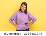 Portrait of young angry serious woman looking at camera with strict negative expression, aggression, keeping hands on hips, wearing purple hoodie. Indoor studio shot isolated on yellow background.