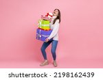 Full length portrait of happy brunette woman holding heavy stack of presents, celebrating holiday, wearing white casual style sweater. Indoor studio shot isolated on pink background.