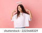 Small photo of Portrait of cute little girl wearing white T-shirt pointing at holy nimbus over head, showing aureole and looking up with toothy smile. Indoor studio shot isolated on pink background.