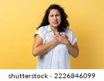 Small photo of Portrait of sick attractive woman with dark wavy hair holding hand on chest, feeling awful acute pain in heart, risk of heart attack. Indoor studio shot isolated on yellow background.