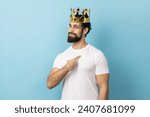 Small photo of Portrait of proud man with beard wearing white T-shirt and golden crown and pointing himself, looking at camera with smile, superior privileged status. Indoor studio shot isolated on blue background.