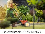 Small photo of Caucasian Professional Gardener with Push Spreader Fertilizing Residential Lawn For a Good Health and Appearance of the Grass. Garden Maintenance.
