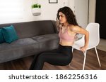 Small photo of Athletic latin woman in her 20s working out her arms with tricep dips exercises. Good-looking woman using a chair for her home workout