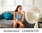 Small photo of Happy woman trying to cool down drinking an ice cold drink and turning on the electric fan at home