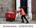 Small photo of WINDSOR, UK - SEPTEMBER 15 2022: Postman in orange jacket employed by England's Royal Mail pushes a large trolley carrying mail through the streets of Windsor, passing a front door