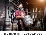 Young male brewer in leather apron holds barrel with craft beer at modern brewery factory