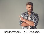 Handsome bearded man in casual clothes is pointing away, looking at camera and smiling, on gray background