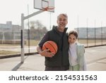 Small photo of Happy father and teen daughter embracing and looking at camera outside at basketball court.