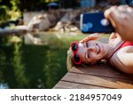 Rlaxed woman relaxing on the pier, lying and taking selfie near the natural lake in summer