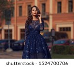 Small photo of Elegant woman in blue ball gown dress standing and posing on a sunny evening at city street. Fashion model full length portrait