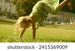 Small photo of Little blonde girl performs handspring on a green park lawn on a sunny summer day
