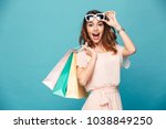 Portrait of an excited beautiful girl wearing dress and sunglasses holding shopping bags isolated over blue background