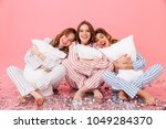 Small photo of Photo of happy women 20s wearing leisure clothings sitting barefoot on floor with legs crossed and hugging pillows during girlish sleepover isolated over pink background