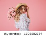 Close up portrait of an attractive young woman in summer dress and straw hat holding carnations bouquet and looking over her shoulder isolated over pink background