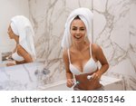Laughing young woman in bra and towel around her head brushing her teeth while leaning on a sink at the bathroom