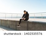 Photo of stylish guy 20s in black tracksuit tying his shoelaces while running by seaside along pier during morning workout