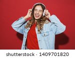 Portrait of a cheerful stylish young woman wearing denim jacket standing isolated over red background, listening to music with headphones, dancing