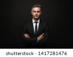 Handsome confident businessman wearing suit standing isolated over black background, arms folded
