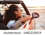 Image of young pretty woman smiling and looking aside while riding in convertible stylish car by seaside