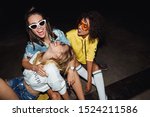 Image of happy multinational girls in streetwear smiling and sitting on skateboards at night party outdoors