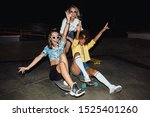 Image of three multinational girls in streetwear smiling and riding on skateboards at night party outdoors