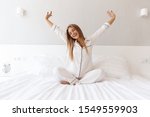 Photo of young happy woman in pajama stretching her arms and smiling while sitting on bed after sleep or nap