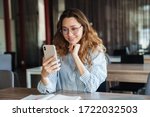 Image of joyful charming woman in eyeglasses smiling and using cellphone while studying at classroom